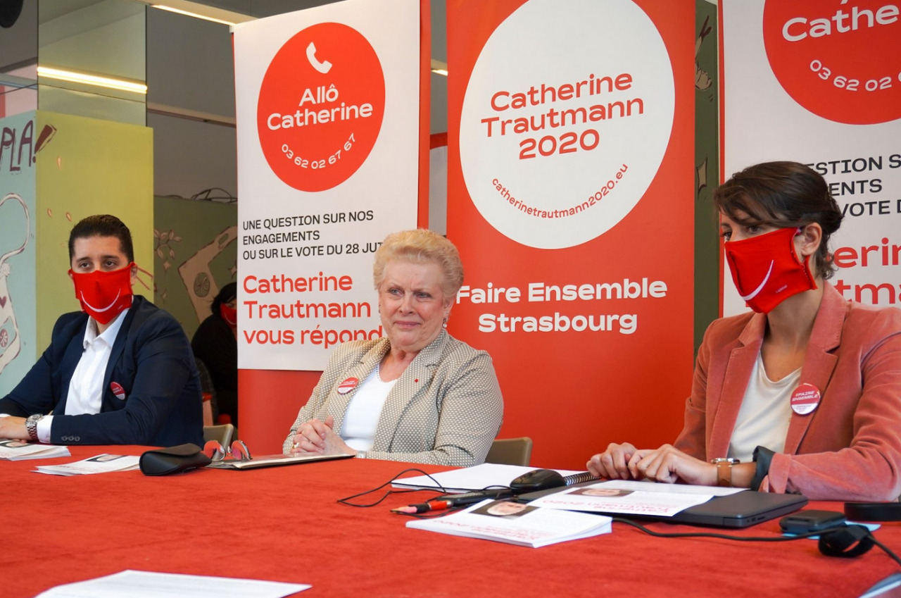 Image 1 : Photographie de Catherine Trautmann en campagne municipale pour la ville de Strasbourg en 2020, entourée de deux personnes portant un masque sanitaire avec un sourire blanc sur fond rouge